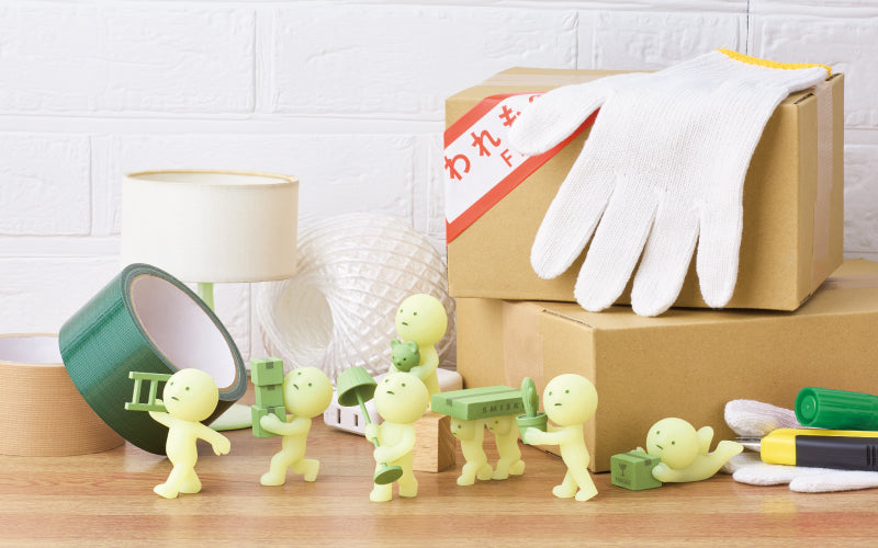 SMISKI Moving Series shows SMISKI minifigures against a while brick wall on top of a wooden table. Green duct tape, a small lamp, white thread, cardboard boxes, gloves and tools surround the SMISKI minifigures. From left to right minifigures include: SMISKI Carrying Ladder, SMISKI Balancing Boxes, SMISKI Decorating, SMISKI Teamwork, Green Thumb SMISKI and SMISKI Falling Down