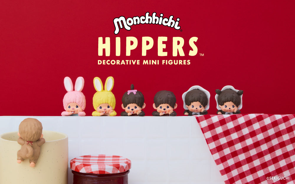 Monchhichi HIPPERS Decorative Mini Figures banner on a red background announces new product. Monchhichi Hippers are in a line; pink bunny, yellow bunny, girl figure, boy figure, baby boy with hood, and baby girl with hood are against a red, white and checkerboard background. A tan figure is set on a mug, facing away next to a jar of red jam
