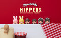 Monchhichi HIPPERS Decorative Mini Figures banner on a red background announces new product. Monchhichi Hippers are in a line; pink bunny, yellow bunny, girl figure, boy figure, baby boy with hood, and baby girl with hood are against a red, white and checkerboard background. A tan figure is set on a mug, facing away next to a jar of red jam