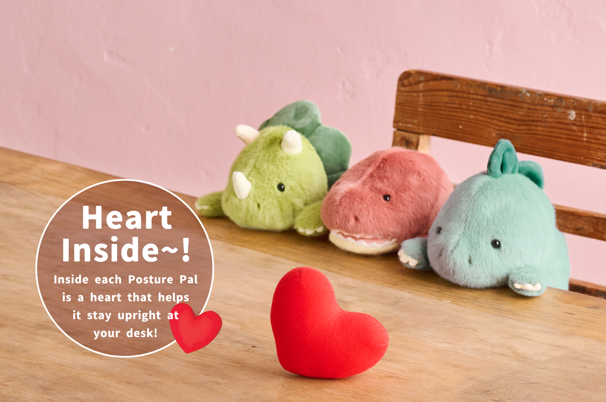 Posture Pal Dinosaurs sit on wooden chair at wooden table against pink background. Left to right: light and dark green Triceratops, light red and white Tyrannosaurus Rex and light and dark teal Stegosaurus. Red heart plush sits on table in front of them. White text in white circle states "Heart inside~! Inside each Posture Pal is a heart that helps it stay upright at your desk!" with a heart icon in bottom edge of circle.