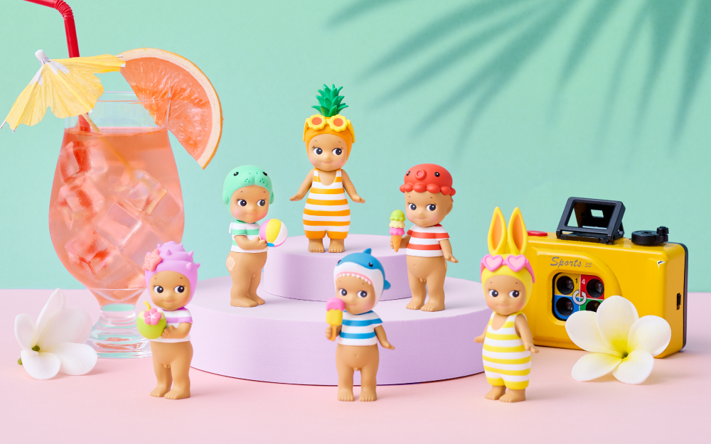 Set against a light teal background, atop a light pink counter top sits a pink drink with fruit and umbrella, gardenia flower blossoms and a yellow point and click camera. Centered in the image is the full line up of the Summer Vacation Series in a pyramid configuration. Atop the pyramid, then left to right: Pineapple, Manatee, Octopus, Shell, Shark and Rabbit minifigures in their white and colored striped swimsuits, holding a variety of summer themed items.