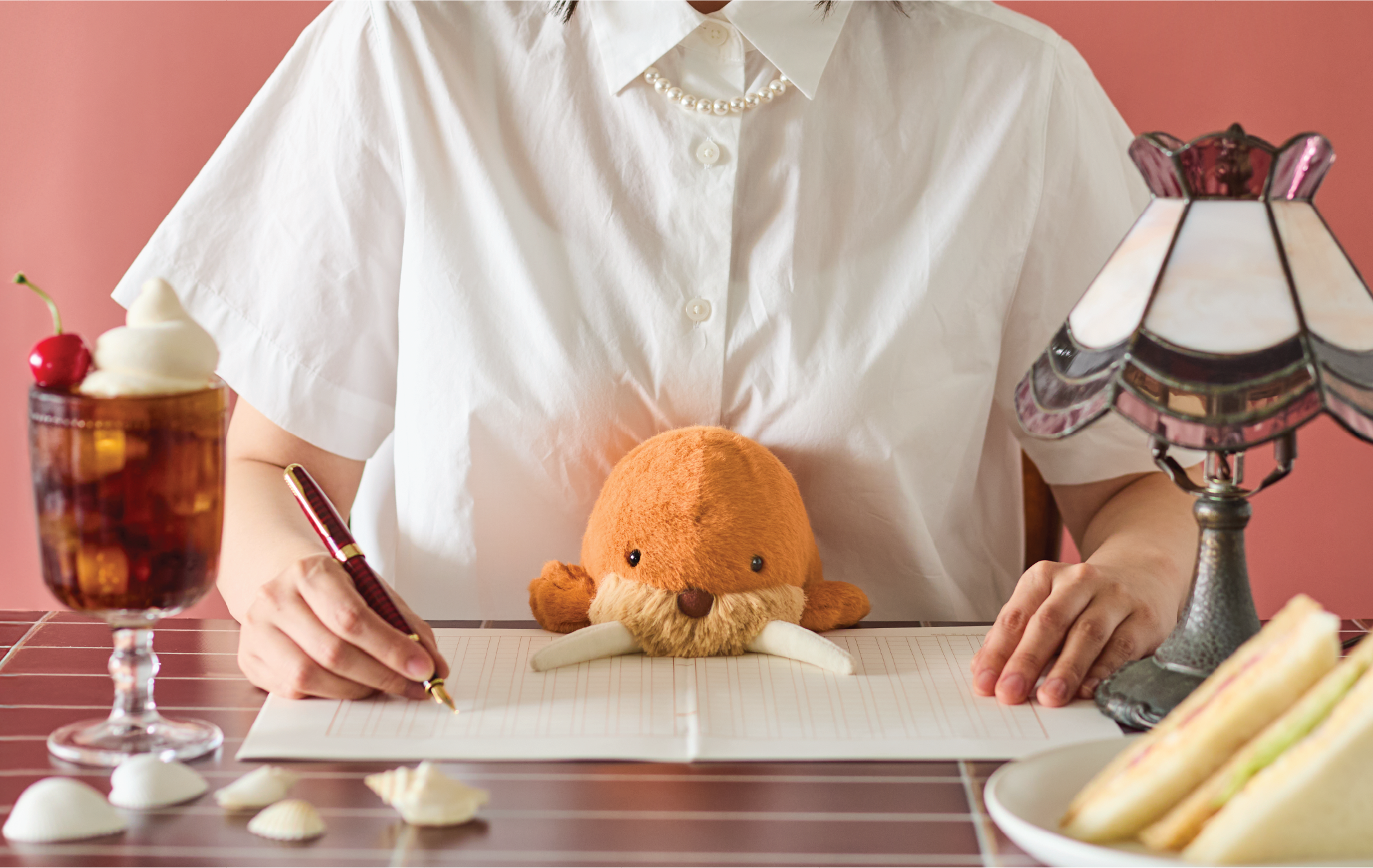 Image shows the upper body of a person in a white button down shirt and pearl necklace sitting with the brown walrus Posture Pal in their lap with a pen in hand, and paper on a wooden table which has a pink and white lamp, sandwich, seashells and soda float on it. the image is set on a dark pink background.