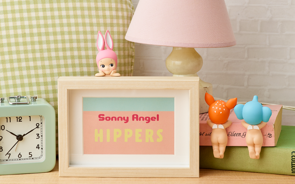 nightstand set with picture frame that reads SONNY ANGEL HIPPERS with sonny angel rabbit hipper attached facing the camera. on the right fawn and elephant are attached to the spine of a book. lamp, pillow, and alarm clock props included.