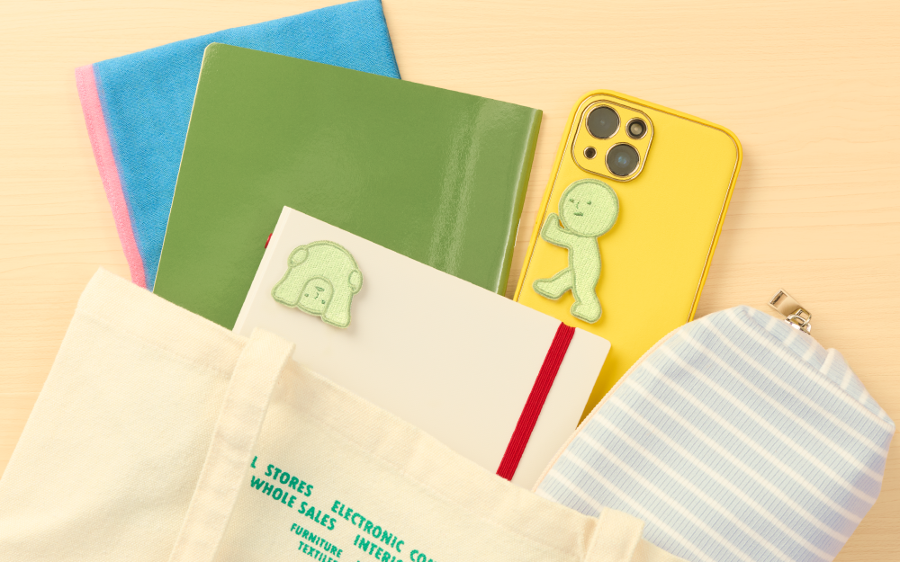 Image set on wooden table background, above canvas tote bag, Peeking sticker variant on upper left corner of white and red notebook, beside yellow phone case with Pushing sticker variant beside green folder, blue folded towel and blue and white zipper bag.
