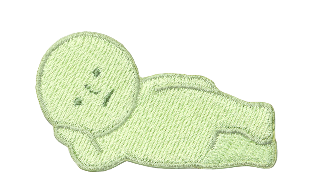 Image with transparent background shows SMISKI Embroidery Sticker "Lounging" SMISKI is a light green figure with dark green eyes, nose and mouth lying on their side, propped up by one hand.