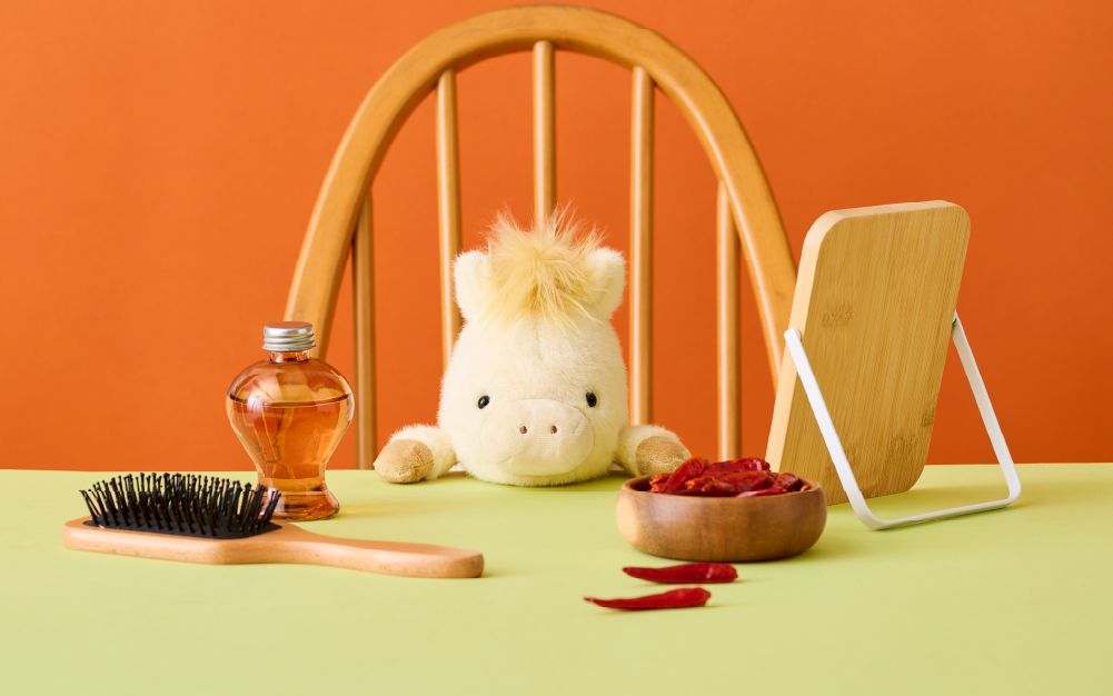 2026 Posture Pal Horse: White product shows the Posture Pal Horse on a wooden chair at a green table top, against a rich orange background. Atop the table top, there is a hairbrush beside a bottle of oil, a small tabletop mirror, and wooden bowl of chili peppers.