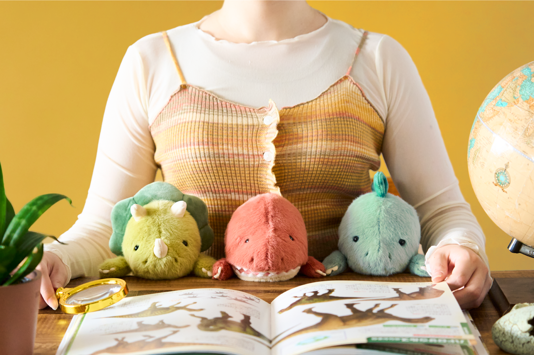 Set on a yellow background, figure in white and yellow striped shirt sits with Posture Pal Dinosaur line up. From left to right, dark and light green Triceratops, light red and white Tyrannosaurus Rex, and light and dark teal Stegosaurus. Figure sits with dinosaur book and magnifying glass beside potted plant, globe and dinosaur egg figurine.
