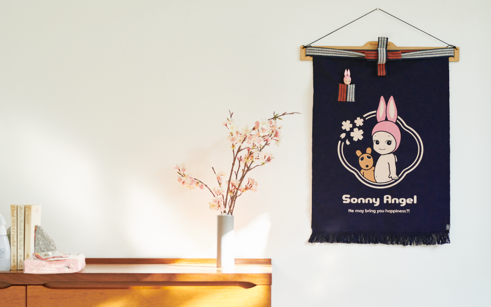 Image set on a white background. A wooden table shows several books, and items beside cherry blossom branches in a white vase. On the right side of image, the Maekake apron is hung on a wooden hanger. Apron's design: Rabbit Sonny Angel beside Robby encircled with cherry blossoms, and text: "Sonny Angel" "He may bring you happiness?!" Rabbit minifigure is tucked into the red and white pocket of apron.