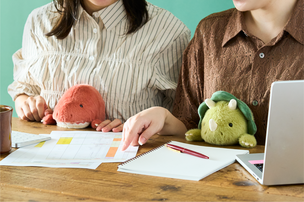 Set on a teal background. Two figures, one in white and gray striped shirt, the other in a brown pattern button down shirt sit side by side at a desk looking over a notebook and papers. A laptop, keyboard and mug are set on the desk with them. On left, light red and white Tyrannosaurus Rex Posture Pal supports one figure, and the other holds the light and dark green Triceratops Posture Pal