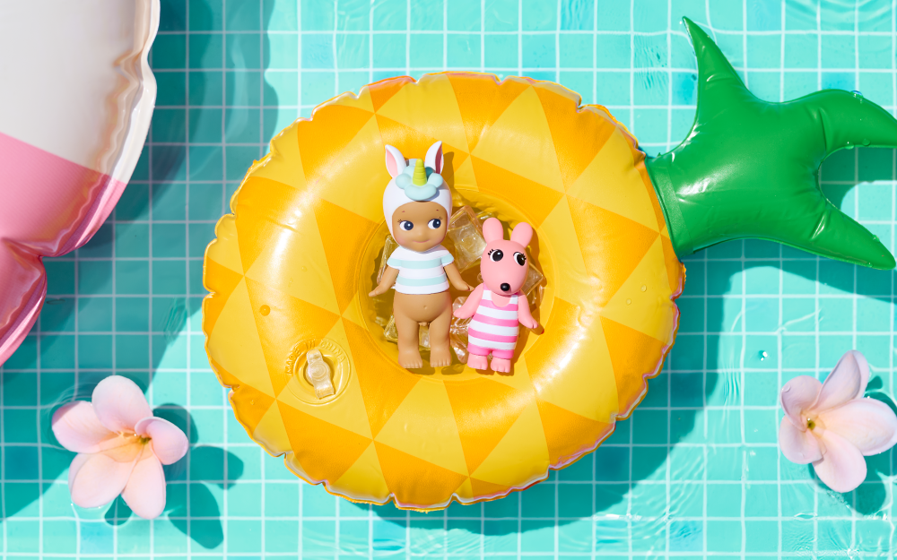Set against a blue tiled background, centered in a pineapple pool float are the secret minifigure and Robby Angel minifigure from the Summer Vacation Series. Secret Sonny Angel wears unicorn headgear and a light blue and white striped shirt. Beside Sonny, Robby Angel is pink with a pink and white striped bathing suit. Flowers float in the water around them.