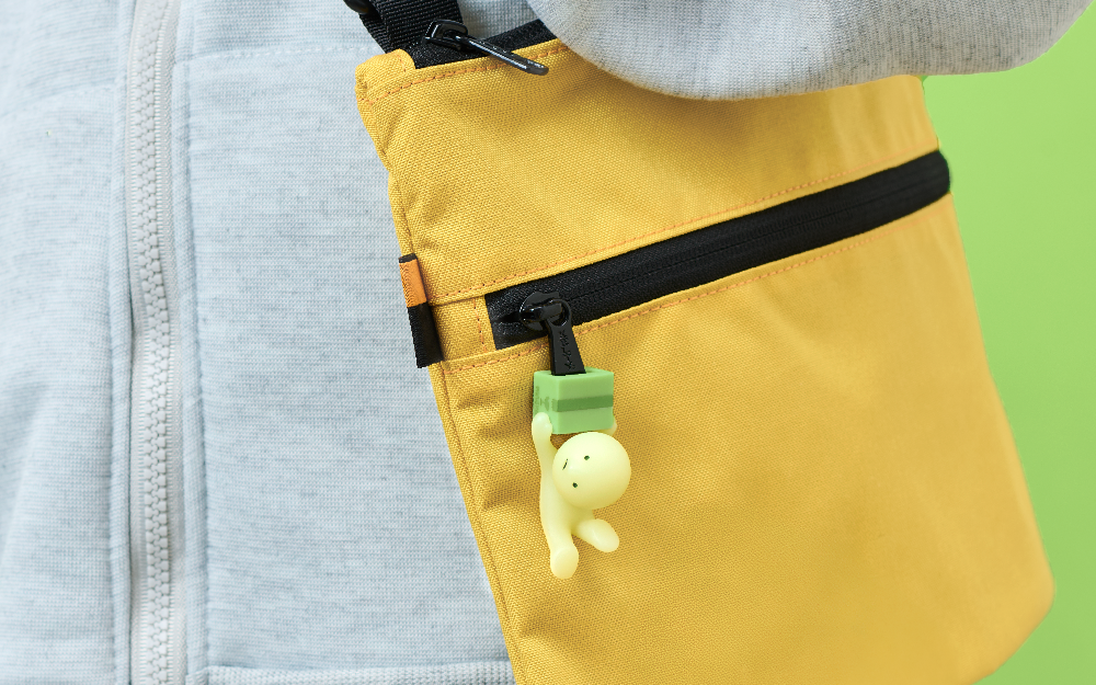 "Falling Down" SMISKI Zipperbite is attached to the black zipper of a black and yellow shoulder bag. The bag is being held by a person in a grey zippered sweatshirt and set on a bright green background.