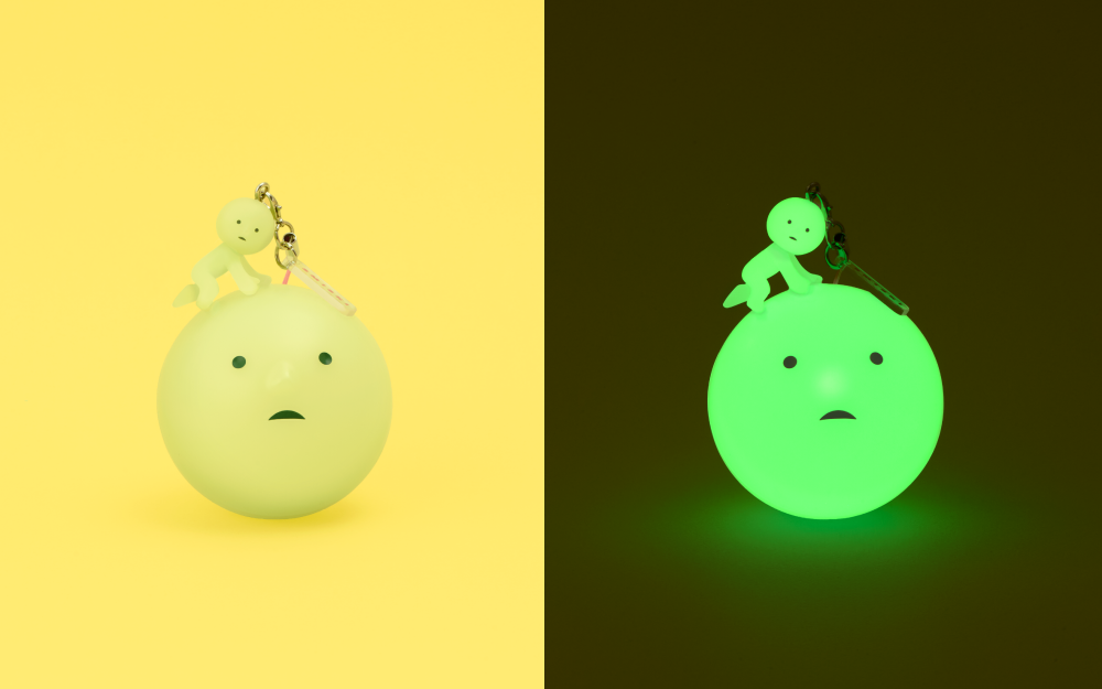 lifestyle image split in half. left: smiski charm balanced on gachapon capsule in light on yellow background. right: same image in the dark showing the SMISKI charm and capsule glowing.