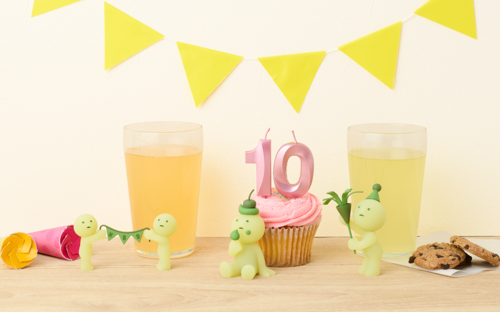 Image set on white background and wooden table, yellow banner set against white wall. Atop wooden table are two glasses of punch, several cookies and party poppers. In the center is a pink frosted cupcake with candles in the shape of the number "10". SMISKIs holding a "HBD" banner, SMISKI sitting with a cherry atop its head eating frosting and SMISKI holding a confetti popper with a party hat atop its head shows off some of the new minifigures.