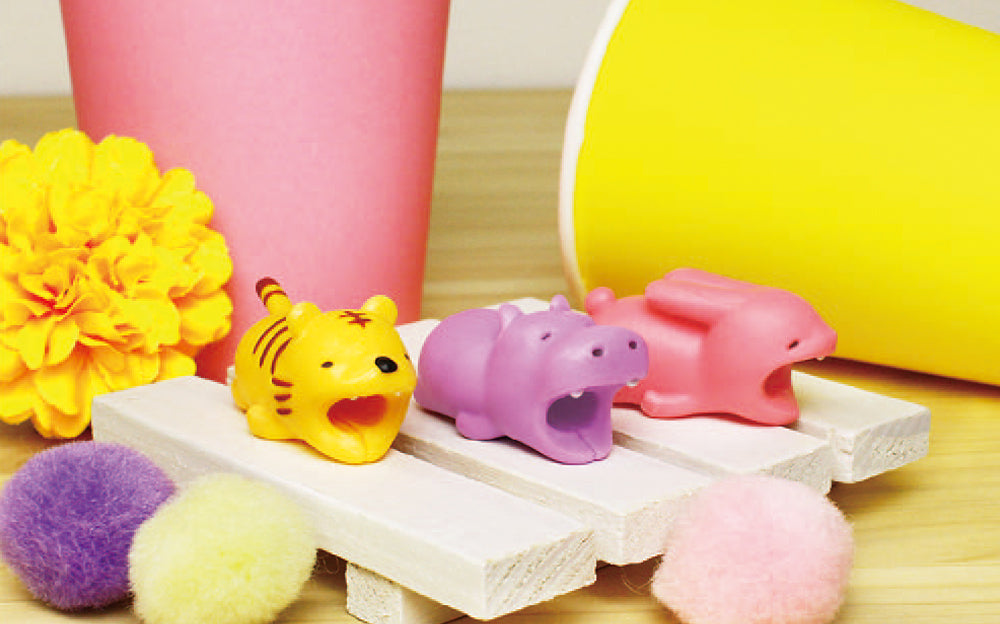 Image shows Cable Bite Vol 1 product variants set against a bright image with yellow, pink and purple pompoms, flowers and cups. Atop a small wooden pallet, left to right: Tiger, Hippopotamus, and Rabbit
